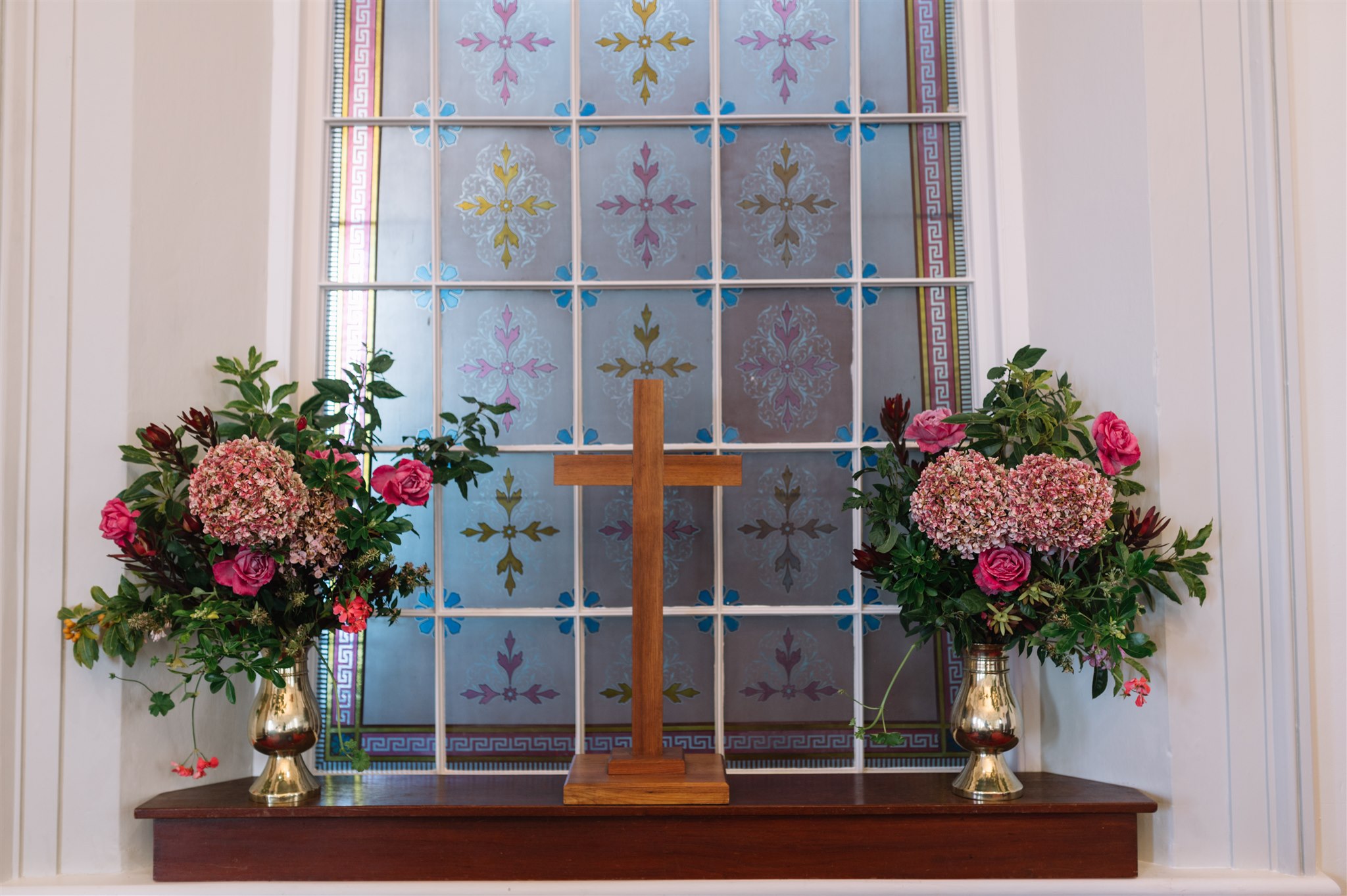 A wooden cross besides vases of fresh flowers in front of a stained glass window